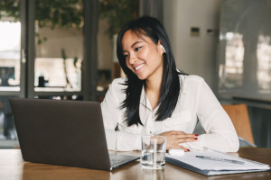 Photo of adorable asian female worker 20s wearing white shirt and earbud smiling while sitting at table in office, and working on laptop